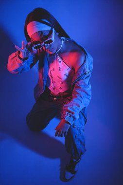 Kafasında güneş gözlüğü olan siyah beyaz elbiseli bir kadın fotoğraf için poz veriyor. Fotoğraf mavi ve mor renkte.