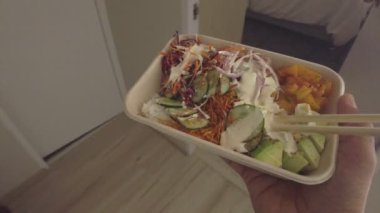 Bir insan elinde yemek çubuklarıyla bir tepsi tutuyor. Tepside havuç, salatalık ve brokoli gibi çeşitli sebzeler yer alıyor. O kişi yemeğinden zevk alıyor gibi görünüyor.