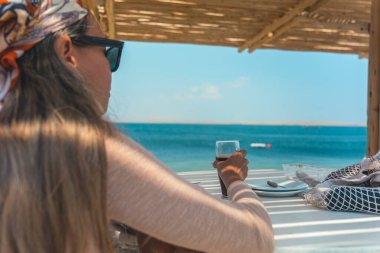 Bir kadın okyanusun kenarında bir masada oturuyor, elinde bir bardak şarap tutuyor. Sahne, arka planda dalgaların sesiyle sakin ve huzurlu.