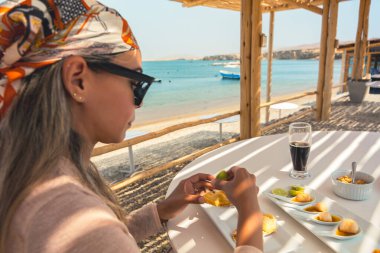 Bir kadın bir bardak bira ve bir tabak yemekle bir masada oturuyor. Güneş gözlüğü ve eşarp takıyor. Sahne, arka planda bir su kütlesiyle bir kumsalda geçiyor.
