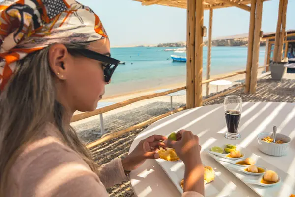 Bir kadın bir bardak bira ve bir tabak yemekle bir masada oturuyor. Güneş gözlüğü ve eşarp takıyor. Sahne, arka planda bir su kütlesiyle bir kumsalda geçiyor.