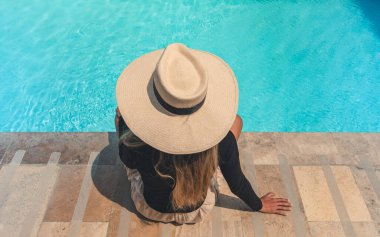 Hasır şapkalı bir kadın havuzun yanındaki taş bir merdivende oturuyor. Şapka büyük ve sarkık, ve kadın suya bakıyor. Sahne huzurlu ve rahatlatıcı.