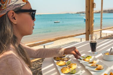 Bir kadın bir bardak bira ve bir tabak yemekle bir masada oturuyor. Masa, bir kase meyve ve bir fincan kahve de dahil olmak üzere çeşitli yiyecek ürünleriyle hazırlandı. Kadın atkı takıyor.