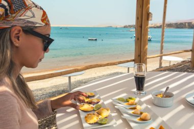 Önünde bir tabak yemekle bir kadın bir masada oturuyor. Güneş gözlüğü ve eşarp takıyor. Masada bir bardak bira ve bir kase yiyecek var. Sahne bir kumsalda geçiyor.