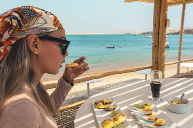 Bir kadın sahilde yemek yiyor. O bir atkı ve güneş gözlüğü takıyor. Masa yiyecek ve içeceklerle dolu.