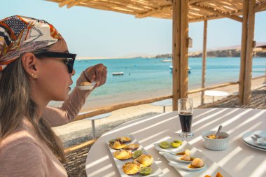 Bir kadın bir bardak bira ve bir tabak yemekle bir masada oturuyor. Masa, bir kase meyve ve bir tabak karides de dahil olmak üzere çeşitli yiyecek ürünleriyle hazırlandı. Kadın atkı takıyor.