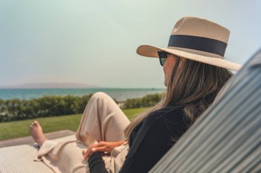 Hasır şapkalı bir kadın okyanusun kenarındaki şezlongda oturuyor. Suya bakıyor ve sahnenin sakin ve huzurlu bir havası var.