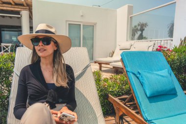 Hasır şapkalı ve güneş gözlüklü bir kadın bahçede bir sandalyeye oturur. Elinde bir cep telefonu tutuyor.