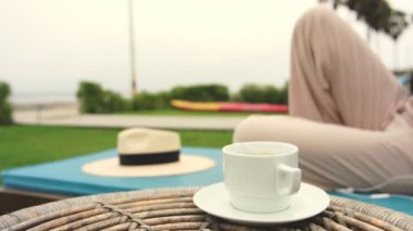 Önünde beyaz bir fincan kahve olan bir kişi katlanır sandalyede oturuyor. Bardağın yanındaki sandalyeye bir hasır şapka yerleştirilir. Sahne, rahat ve sakin bir atmosfer taşır.