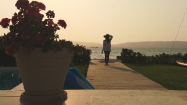 Bir kadın bir su kütlesinin yanında tahta bir tahta kaldırımda yürüyor. Gökyüzü turuncu ve pembe renklerin bir karışımıdır. Huzurlu ve huzurlu bir atmosfer yaratır. Bir saksı bitkisi bir çıkıntıya yerleştirilir.