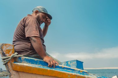 Bir adam başı eğik bir şekilde bir teknede oturuyor. Gökyüzü mavi ve su sakin.