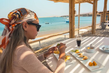 Önünde bir tabak yemekle bir kadın bir masada oturuyor. Turuncu bir eşarp ve güneş gözlüğü takıyor. Masada bir bardak soda ve bir kase meyve var.