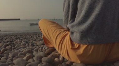 Sahilde arka planda teknesi olan biri oturuyor. Kumsal kayalarla kaplı ve kişi sarı bir etek giyiyor.