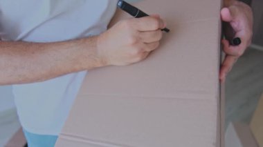 Bir adam kutuları topluyor ve üzerine giysiler yazıyor. Kutular üst üste istiflenmiş ve adam onlardan birine uzanıyor.