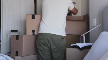 Kutular ve kıyafetlerle dolu bir odada bir adam duruyor. Raftaki bir kutuya uzanıyor.