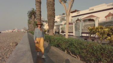 Bir kadın kaldırımda bir evin yanında yürüyor. Ev beyaz ve etrafında bir çit var. Kadın mavi gömlek ve sarı pantolon giyiyor.