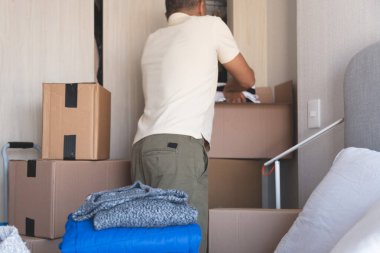 Kutular ve kıyafetlerle dolu bir odada bir adam duruyor. Raftaki bir kutuya uzanıyor.