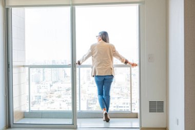 Bir kadın balkonda dikilip şehre bakıyor. Beyaz bir ceket ve kot pantolon giyiyor.