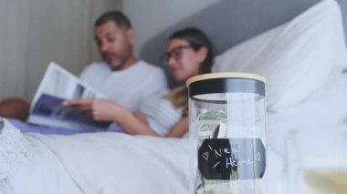 Bir erkek ve bir kadın yatakta uzanmış kitap okuyorlar. Yataklarının yanında bir kavanoz para duruyor.