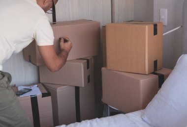 Bir adam keçeli kalemle bir kutuya yazı yazıyor. Kutular üst üste istiflenmiş.