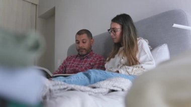 Bir adam ve bir kadın önlerinde bir dergi ile bir yatakta oturuyorlar. Adam dergideki bir şeyi işaret ediyor ve kadın da telefonuna bakıyor. Sahne rahat ve sıradan.