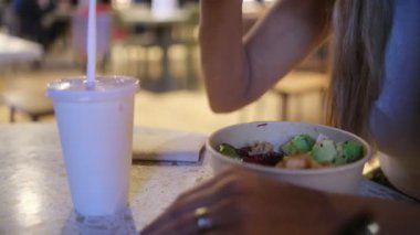 Bir kadın yemek çubuklarıyla salata yiyor ve plastik bardaktan içiyor. Sahne rahat ve rahat, kadın rahat bir ortamda yemeğin tadını çıkarıyor.