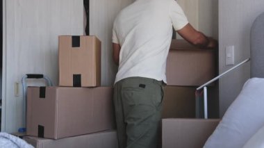 Kutular ve kıyafetlerle dolu bir odada bir adam duruyor. Raftaki bir kutuya uzanıyor.