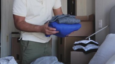 Kutular ve kıyafetlerle dolu bir odada bir adam duruyor. Raftaki bir kutuya uzanıyor.
