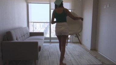 Oturma odasında elinde beyaz bir kutu tutan bir kadın var. Oda seyrek döşenmiş bir kanepe ve bir sandalye ile. Kadın odaya bir şey taşıyor.