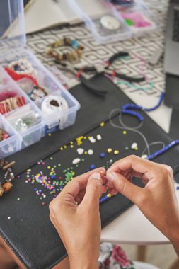 Küçük işletmesi için el yapımı aksesuar koleksiyonu tasarlayan bir kuyumcunun genel görünüşü. Çalışma alanı boncuklarla, aletlerle ve aksesuarlarla dolu. Bu da zanaatkârların yaratıcılığını gösteriyor..