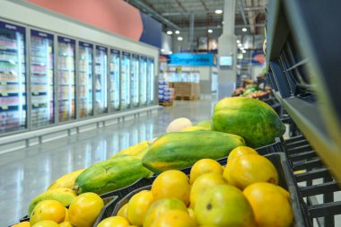 Siyah raflarda taze meyve sergilenen toptan bir süpermarket manzarası. Mango, ananas, papaya ve diğer tropikal ürünler.