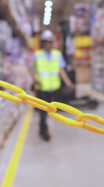 Güvenlik zincirinin odak noktası koruyucu ekipmanlı bir depo işçisi arka planda yürürken, işyeri güvenliğini vurguluyor