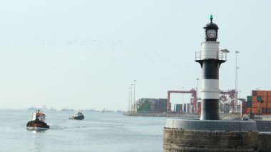 Sahile yakın manzaralı bir deniz feneri, sakin sularda yüzen küçük bir zanaatkar botu ve arka planda liman.