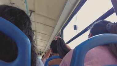 Hareket halindeki bir otobüste oturan uzun siyah saçlı kadın diğer yolculara bakıyor.