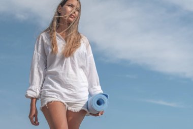 Plajda çıplak ayakla yürüyen genç bir kadın bir yoga minderi taşıyor, meditasyona hazır ve yazın tadını çıkarıyor.