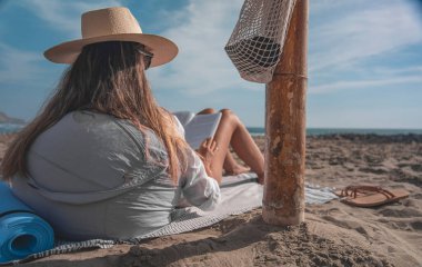 Hasır şapka takan genç bir kadın kumsalda kitap okuyarak yaz meditasyonunun tadını çıkarıyor.