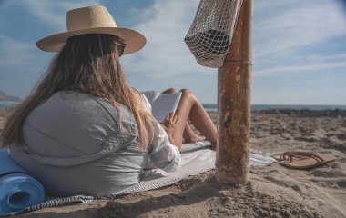 Hasır şapka takan genç bir kadın kumsalda kitap okuyarak yaz meditasyonunun tadını çıkarıyor.
