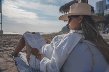 Yaz tatili boyunca güneşli bir sahilde kitap okuyan genç bir kadın.