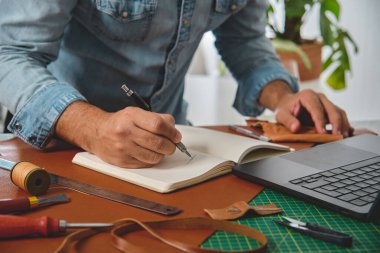 Ayakkabıcı, deri el işi aletleri ve dizüstü bilgisayarla çevrili bir defterde yeni bir model çiziyor.