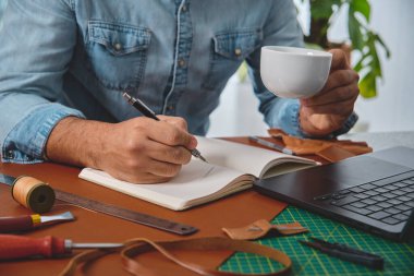 Deri ustası kahve içiyor ve atölyesindeki deftere notlar yazıyor.