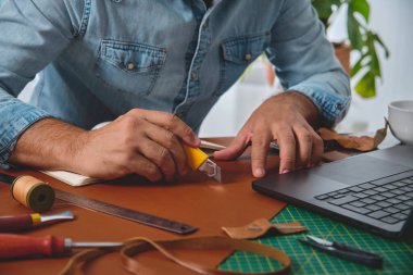Shoemaker atölyesinde dizüstü bilgisayarının yanında deri kesiyor.