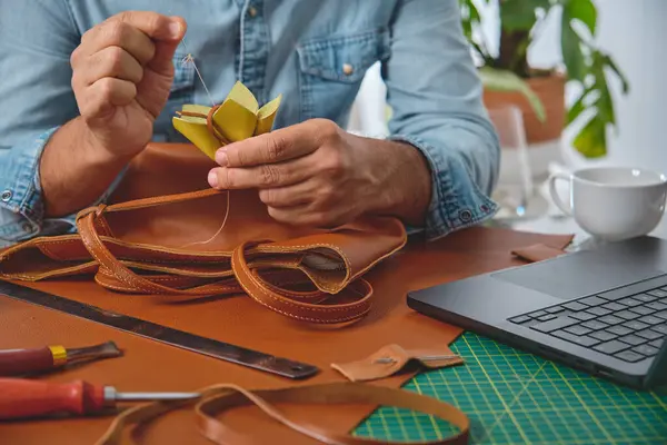 Deri ustası atölyesinde dikkatlice el yapımı bir çanta dikiyor. Etrafı alet ve teknolojiyle çevrili.