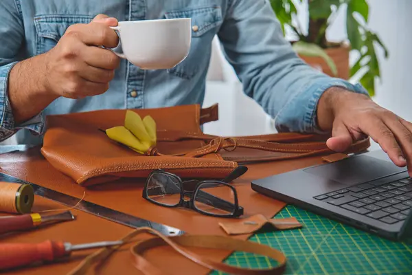 Deri ustası kahve içiyor ve atölyesinde deri işleme aletleriyle dizüstü bilgisayar kullanıyor.