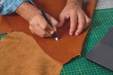 Zanaatkar atölyesinde gümüş bir kalem, dizüstü bilgisayar ve paspas kullanarak deri işaretliyor.