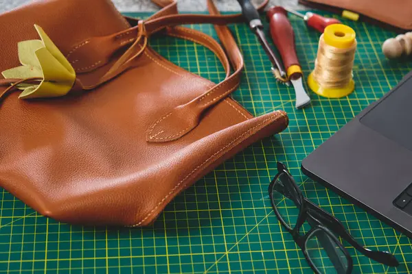 Aletler ve teknolojiyle çevrili küçük bir atölyede el yapımı deri ürünleri sergileyen deri bir el çantası.