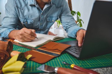 Deri ustası not alıyor ve atölyesinde bir dizüstü bilgisayar üzerinde çalışıyor.