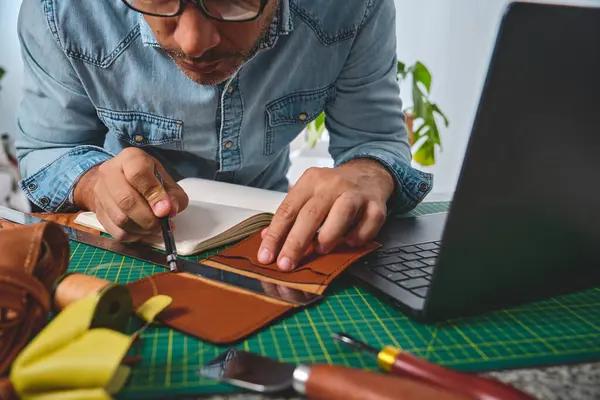 Deri ustası, aletler ve dizüstü bilgisayarla çalışıyor, atölyesinde yeni cüzdan tasarlıyor.