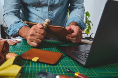 Deri işçiliği yapan zanaatkar elleri küçük işletme atölyesinde dizüstü bilgisayarla el yapımı ürün yaratıyor.