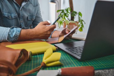Deri ustası atölyesinde online alışveriş için akıllı telefon ve kredi kartı kullanıyor.