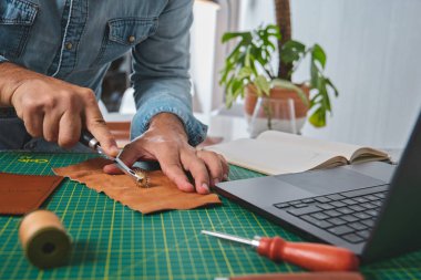 Zanaatkar, atölyesinde dizüstü bilgisayar, defter ve aletlerle dikiş tekerleği kullanarak deri üzerinde çalışıyor.